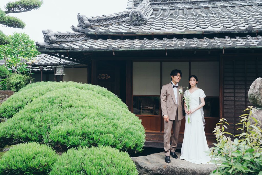 瓦屋根の古民家と緑の庭に立つ新郎新婦