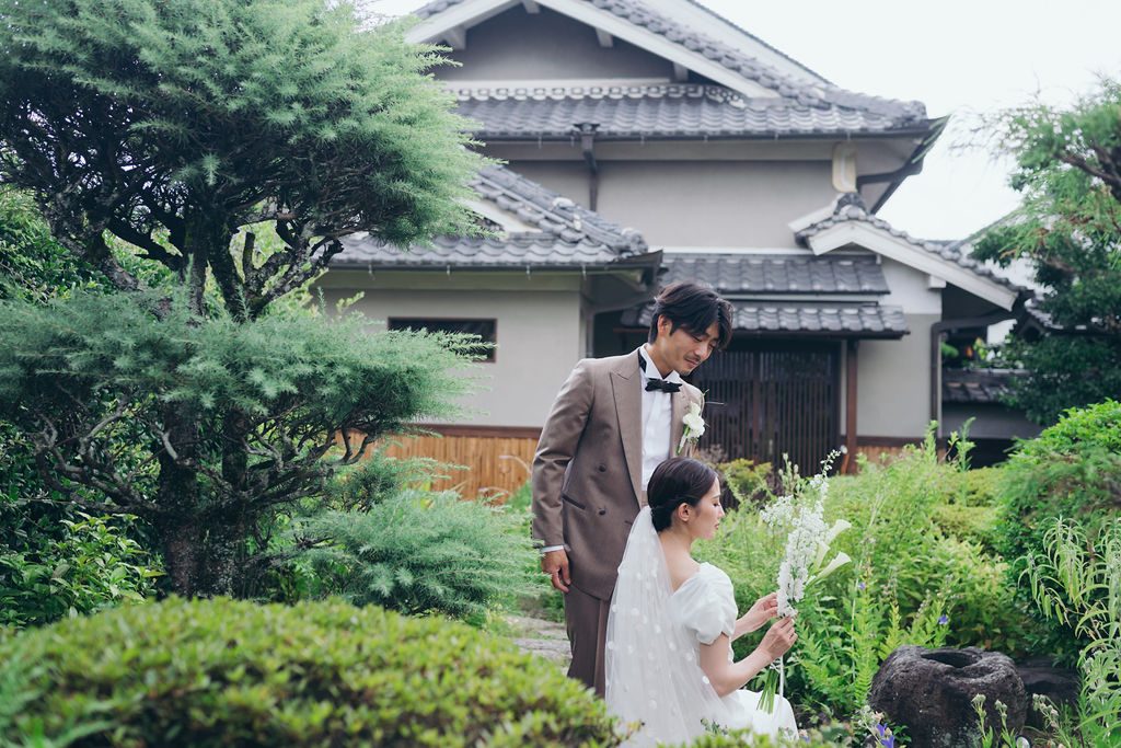 庭で花を手に立つ新郎新婦、瓦屋根を背景にしたロングショット